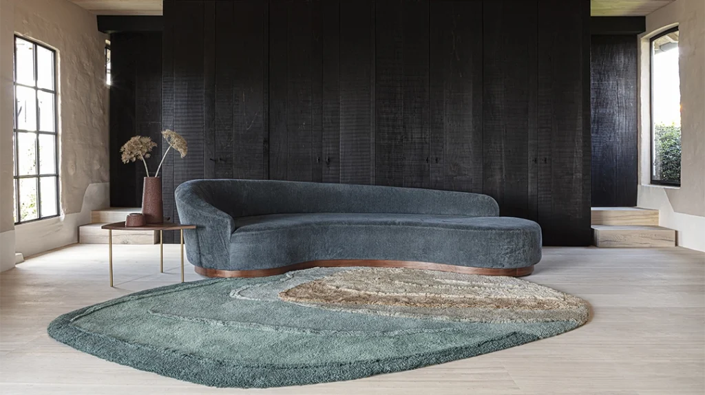 A modern living room with a curved dark teal sofa, an abstract green and beige rug, a small wooden side table, and a vase with dried plants against a black wood-panelled wall and light wooden floors.