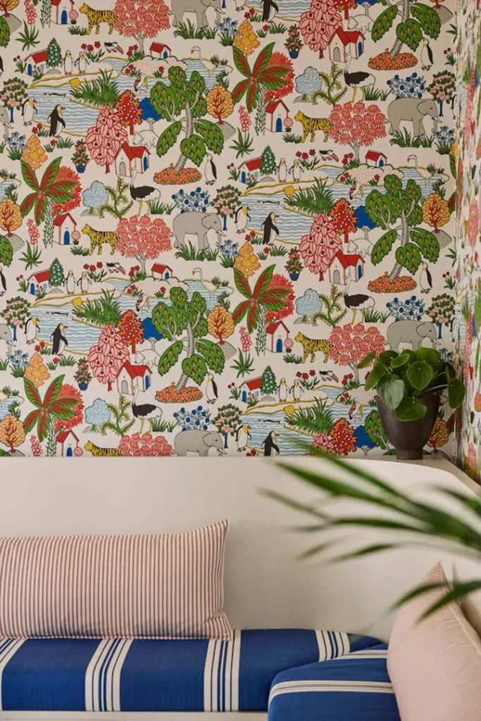 A colourful, patterned wallpaper with illustrated animals, trees, rivers, and houses decorates a wall behind a white sofa. The sofa has striped cushions in red and blue, and a potted plant sits on a ledge.