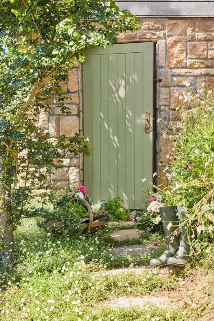 A pale green wooden door set in a stone wall is surrounded by lush garden plants, flowers, and a tree. Green wellington boots and a basket with flowers rest on the stone path leading to the door.