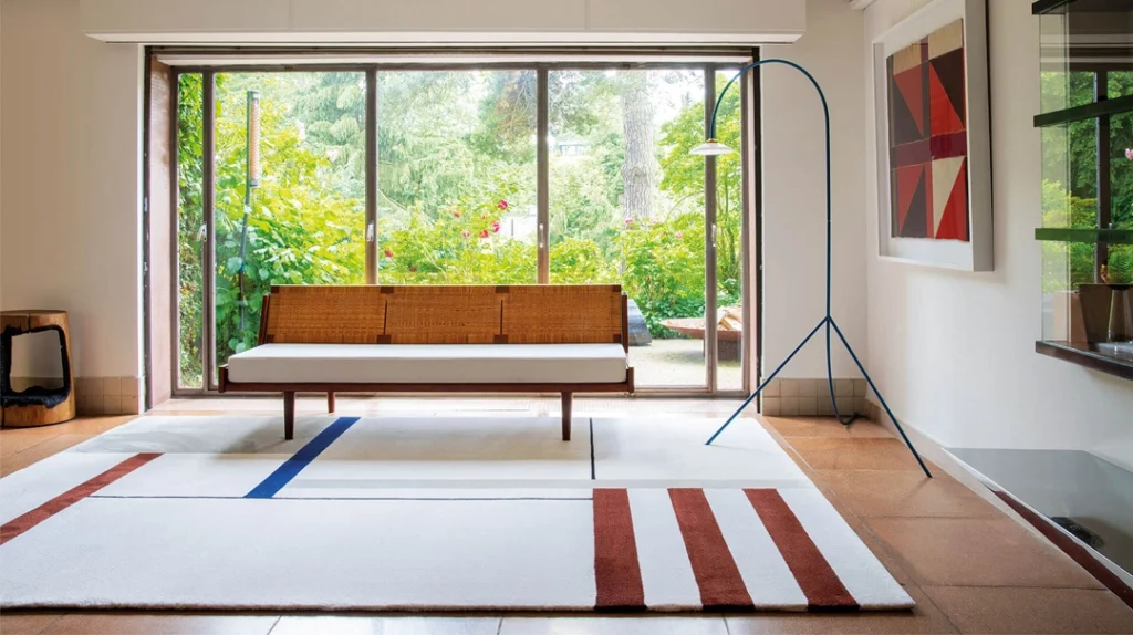 A modern living room with a wooden bench, white cushions, a geometric rug, a tall floor lamp, and large glass doors opening to a lush green garden. Abstract artwork hangs on the wall.