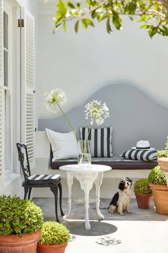 A sunny patio with potted plants, a black-and-white chair, a round white table with a vase of white flowers, a cushioned bench with striped cushions, and a small dog sitting on the ground.