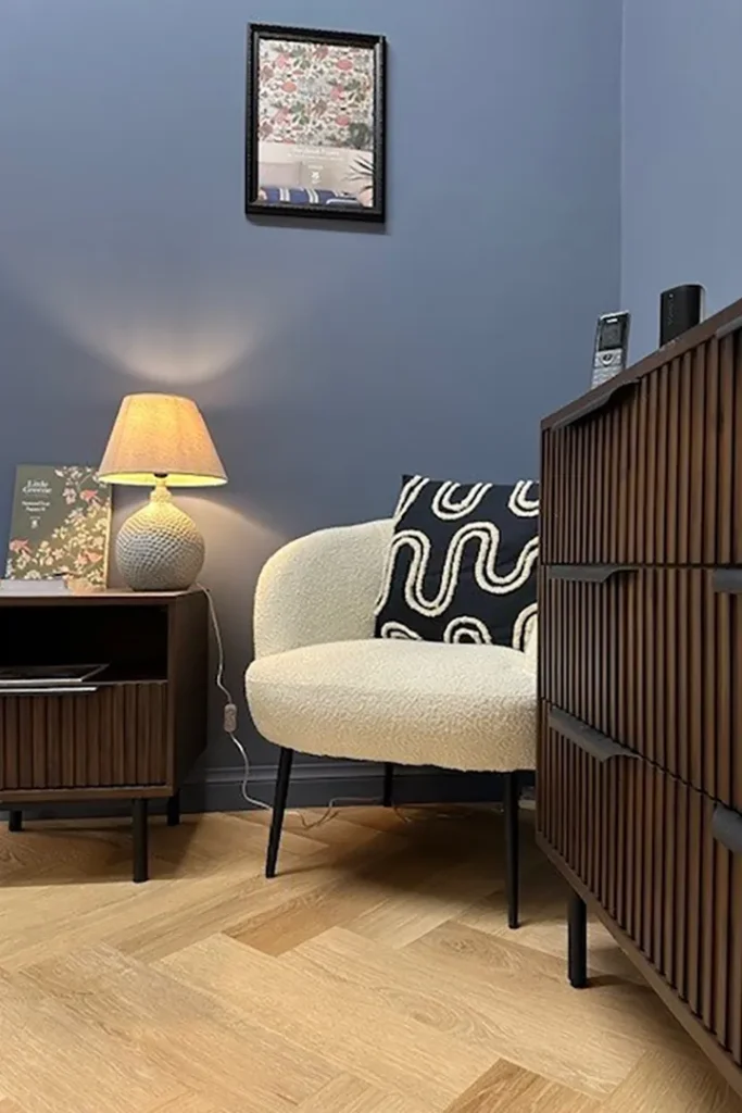 A cosy corner with a cream armchair, black and white patterned cushion, wooden furniture, and a lit table lamp against blue walls and a light wood floor. A framed picture hangs above the chair.