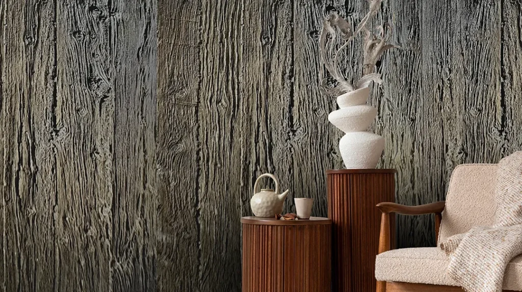 A cosy chair sits beside two wooden side tables holding a teapot, cup, and a stacked white vase with branches, all set against a textured wall that resembles rough tree bark.