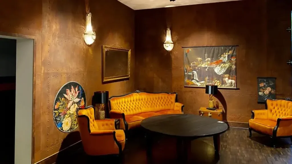 A stylish room with brown walls, an orange tufted sofa, two matching armchairs, a round black table, decorative wall art, and two wall sconces providing warm lighting.