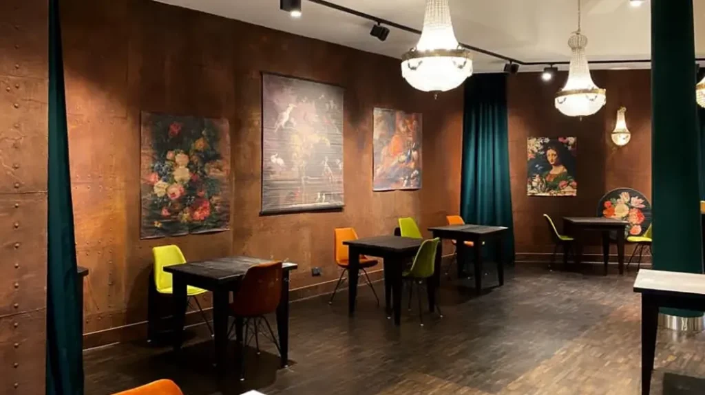 A cosy café with dark wooden floors, small black tables, and colourful chairs. Chandeliers hang from the ceiling, and classic floral and portrait paintings decorate brown walls. Teal curtains hang at the corners.