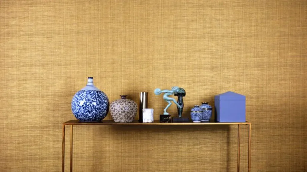 A gold console table holds blue and white ceramic vases, a metallic vase, a green sculpture, and a blue box against a textured gold wall background.