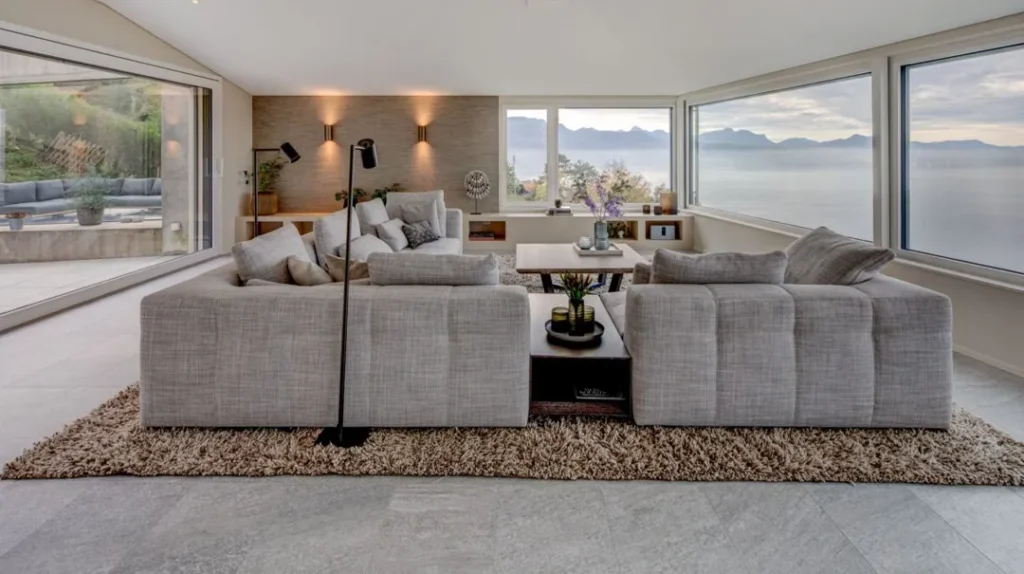 Modern living room with two grey sofas facing each other on a beige rug, large windows offering scenic views of water and mountains, and neutral-toned decor with wall lights and plants.