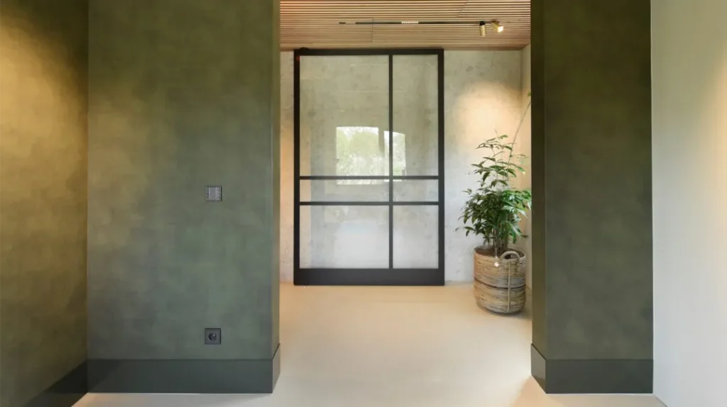 A modern hallway with green-textured walls, a light floor, and a potted plant next to a large glass sliding door featuring black metal frames. Natural light enters from outside, creating a bright atmosphere.