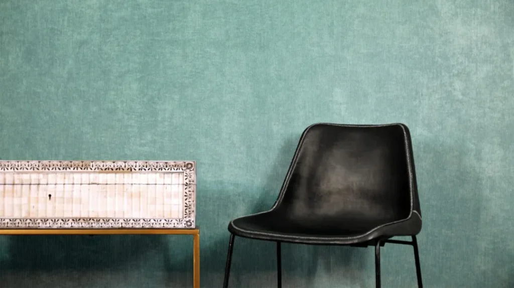 A black modern chair sits next to a patterned box on a wooden table, against a textured teal wall background.