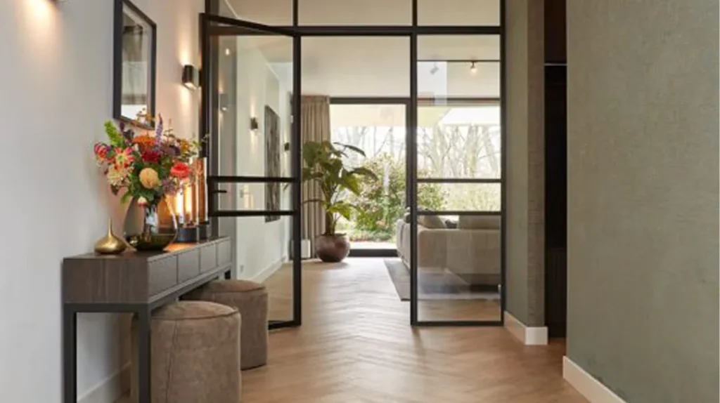 Modern hallway with wooden herringbone floor, a console table with flowers and candles, two round stools, and glass doors leading to a bright living area with large plants and outdoor views.