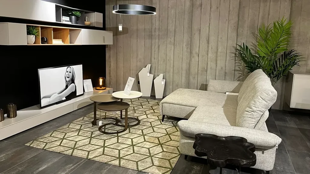 Modern living room with a grey sectional sofa, geometric rug, nested round coffee tables, TV, shelves with decor, potted plant, and wooden wall panels, all in a cosy, stylish setting with soft lighting.