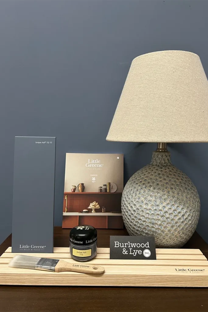 A table displays a decorative lamp, a paint swatch, a Little Greene paint tin and brush, a colour brochure, a Burlwood & Lye business card, and a wooden board, all set against a blue wall.