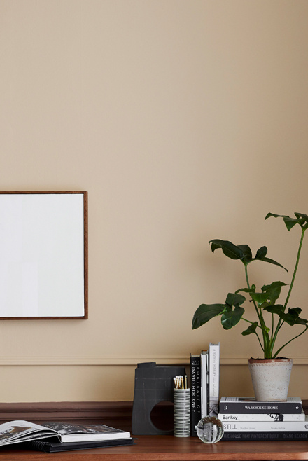 A minimal desk setup with books, stationery, an open magazine, a potted plant, and a blank framed picture on a beige wall. The scene is simple and tidy, with neutral and natural tones.