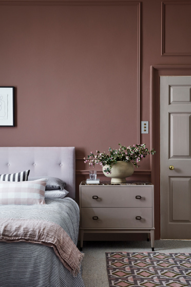 A cosy bedroom with mauve walls, a tufted light grey headboard, a neatly made bed with layered pillows and blankets, a beige chest of drawers with a vase of flowers, and a patterned rug on the floor.