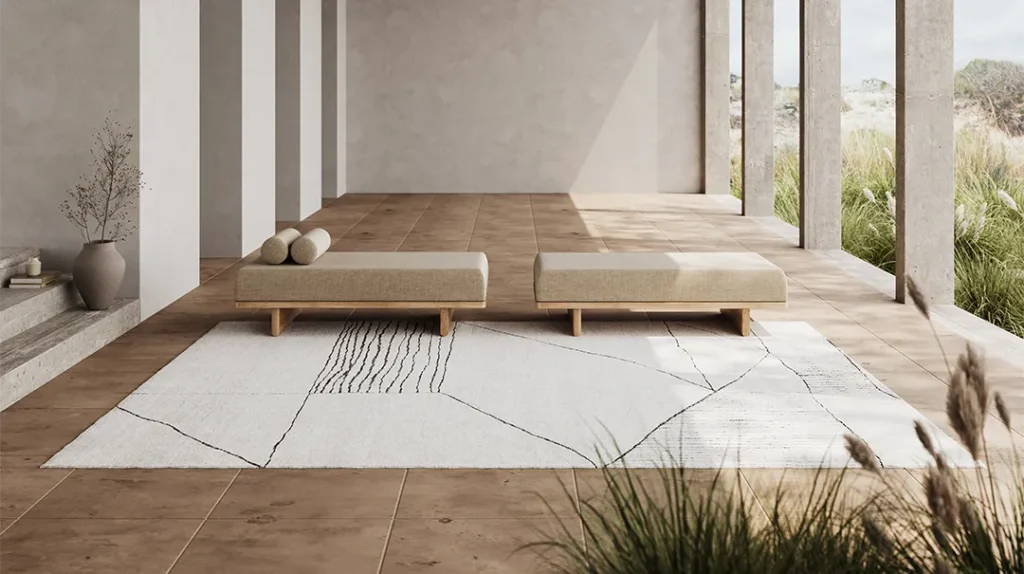 Minimalist patio with two beige cushioned benches on a large white rug with black lines, neutral-toned tiled flooring, vases with dried branches, and open columns overlooking greenery.