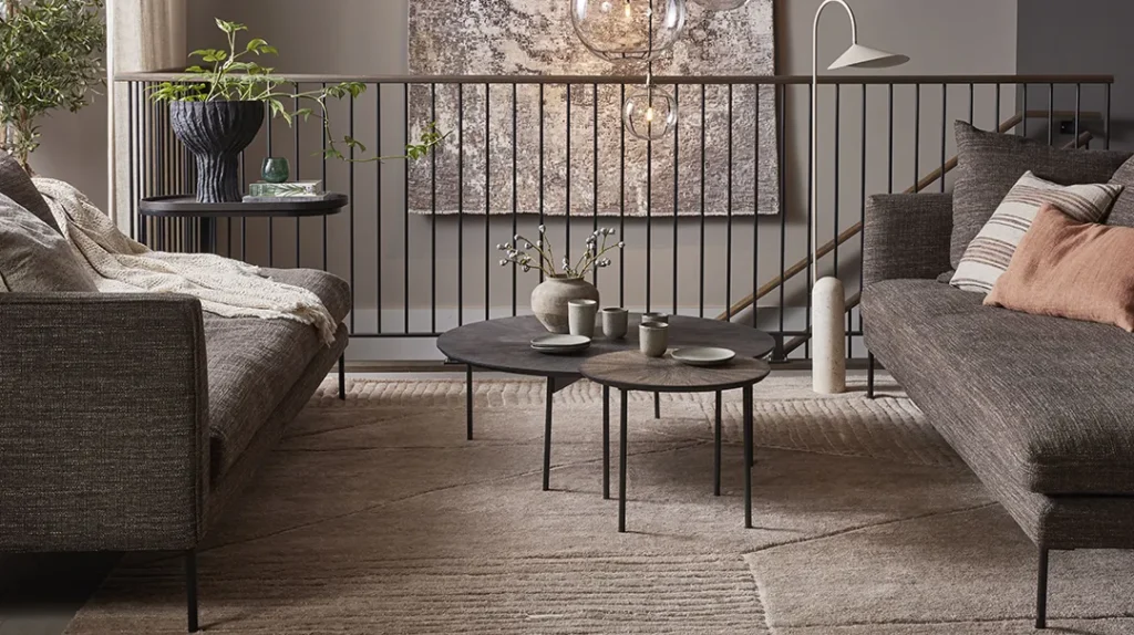 Modern living room with two grey sofas, a layered rug, round black coffee tables, scatter cushions, and plants. Soft lighting and neutral tones create a cosy, inviting atmosphere. A railing and abstract wall art are in the background.