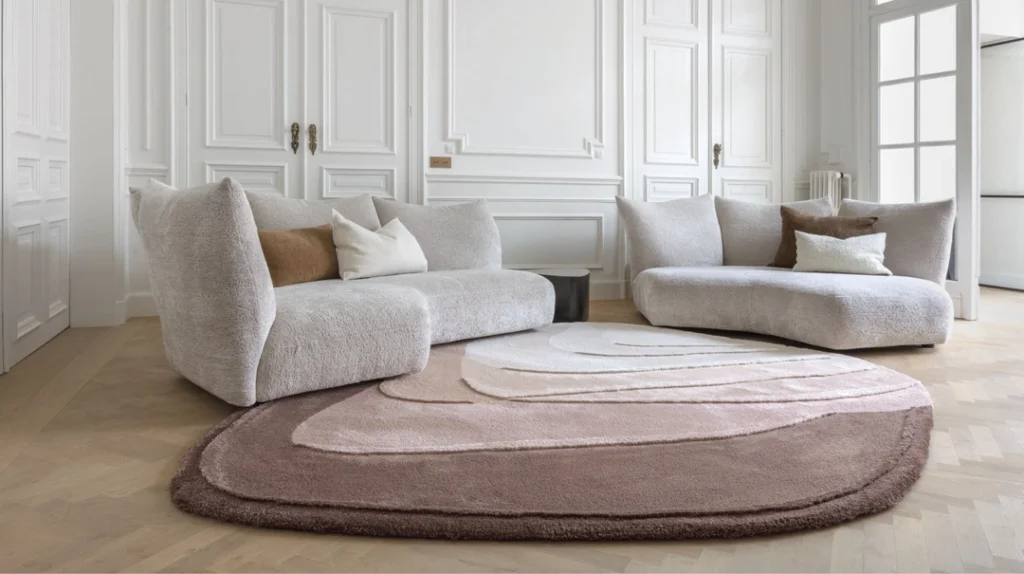 Two modern, light-grey sofas with decorative cushions sit on a large, abstract pink and beige area rug in a bright room with white panelled walls, parquet wood floors, and tall double doors.