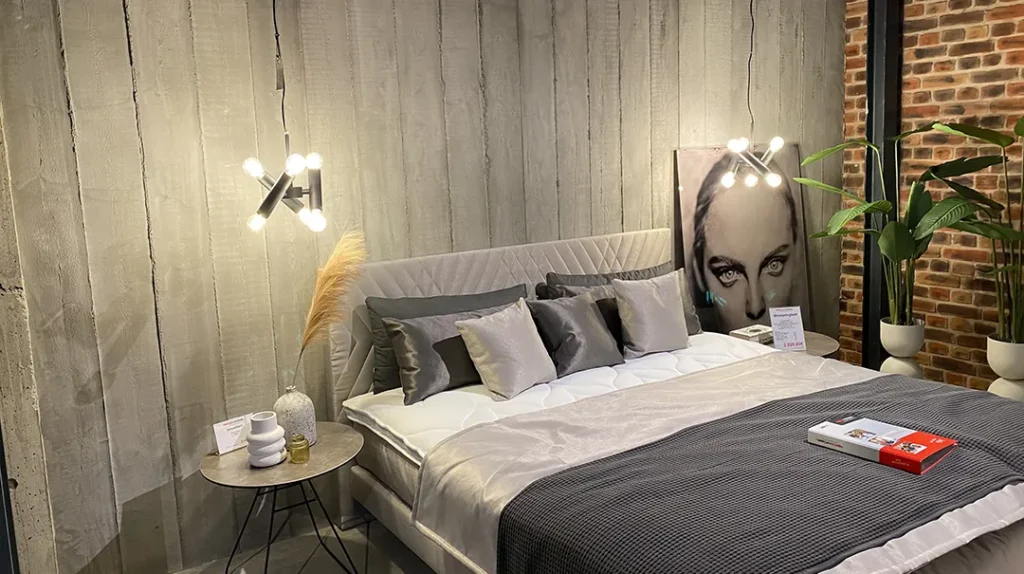 Modern bedroom with a large bed, neutral bedding, two bedside tables, stylish lighting, a plant, and wall art featuring a woman's face. The room has a mix of grey concrete and exposed brick walls.