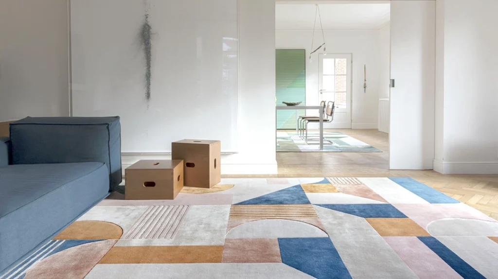 A modern living room with geometric-patterned rug, blue sofa, two wooden cube stools, minimalist white walls, and a view into a bright dining area with green accents and large windows.