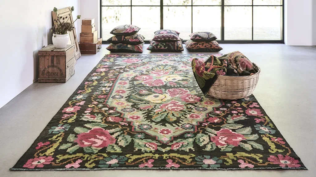 A room with a large floral-patterned rug, colourful cushions stacked by large windows, a basket with folded textiles, and rustic wooden boxes and a bench with a vase of dried flowers in the corner.
