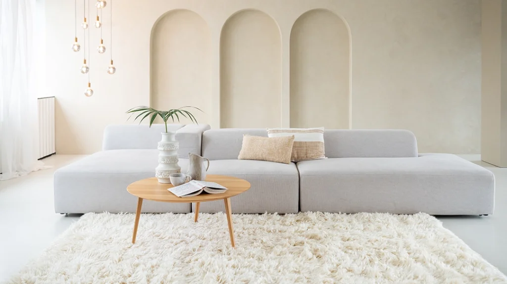 A modern living room with a light grey sofa, a round wooden coffee table with vases and a book, a white shaggy rug, arched wall niches, and hanging pendant lights in a bright, minimalist setting.
