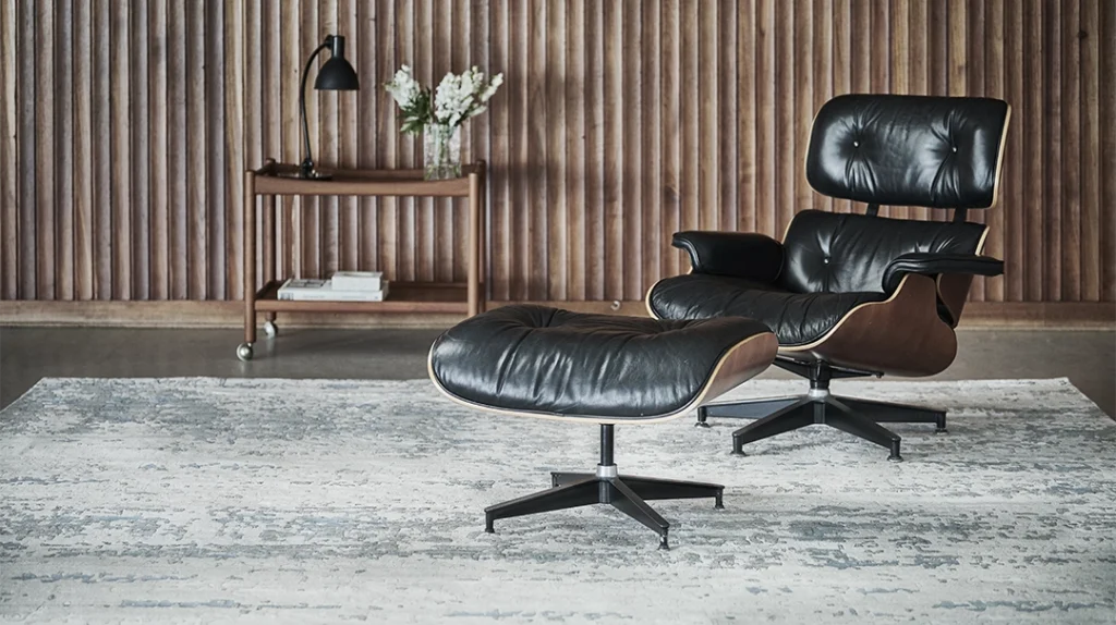 A black leather lounge chair with matching ottoman sits on a light patterned rug in front of a wooden slat wall; a small wooden table with a lamp, flowers, and books is in the background.