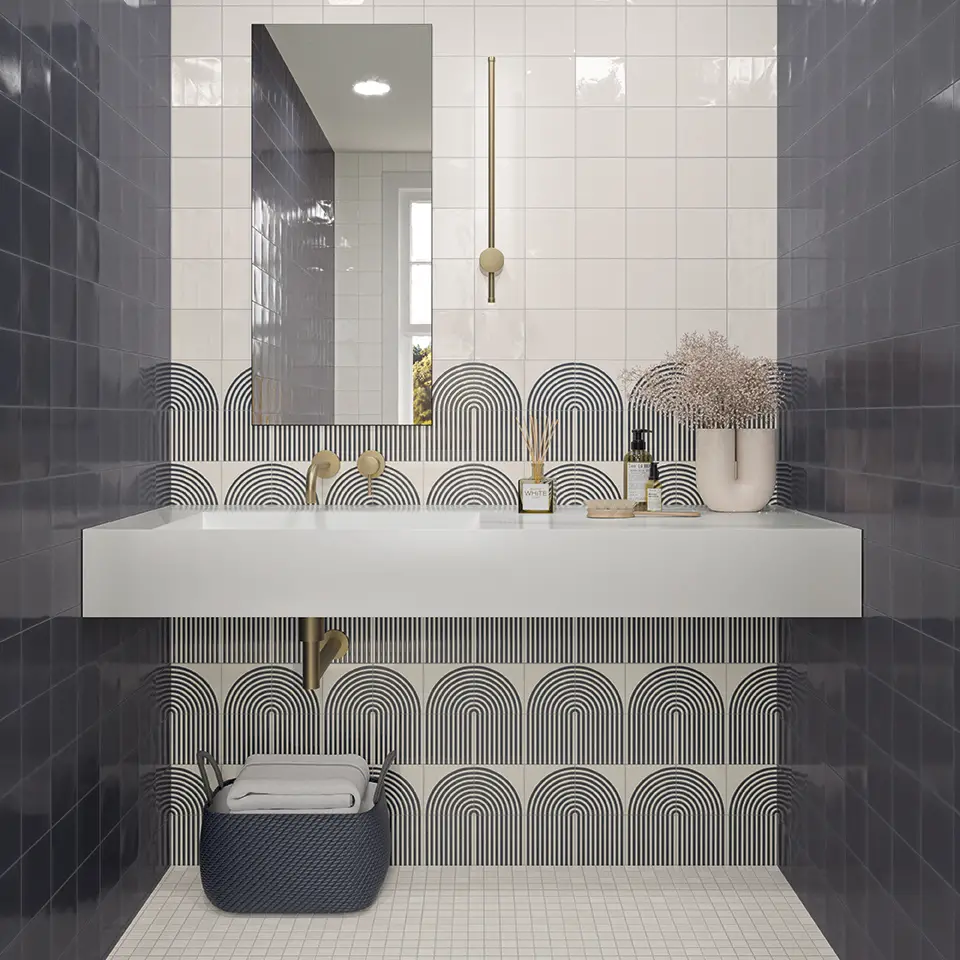 flooring design studio in Chelmsford. Modern bathroom with navy and white geometric tiles, a white floating basin, gold fittings, a rectangular mirror, a hanging pendant light, and a dark basket with towels beneath the basin. A vase with dried flowers sits on the counter.