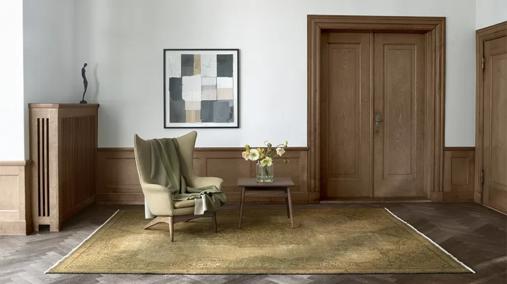 A cosy room with wood-panelled walls and double wooden doors, featuring an armchair draped with a green throw, a small table with a vase of flowers, an abstract painting, and a patterned rug on a herringbone floor.