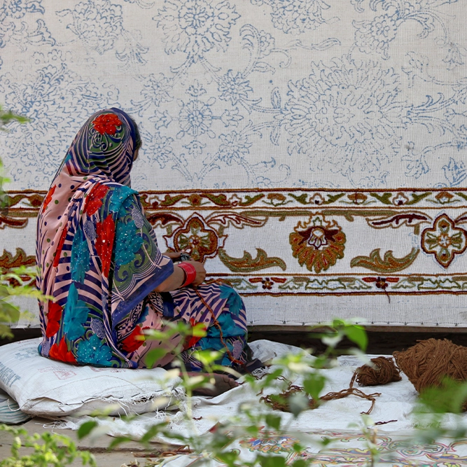 A woman in colourful traditional clothing sits on cushions, weaving an intricate, patterned textile with floral designs. Yarn and materials are scattered nearby; green plants frame the scene in the foreground.