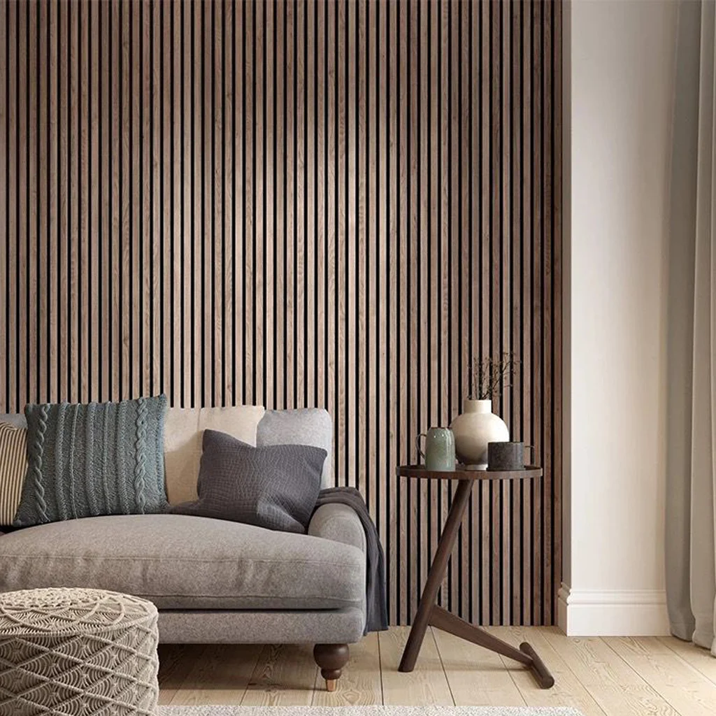 A modern living room with a beige chaise longue, grey and green cushions, a round side table with decorative vases, and a wooden slat feature wall. Light curtains hang to the right, and a woven pouffe sits in the foreground.