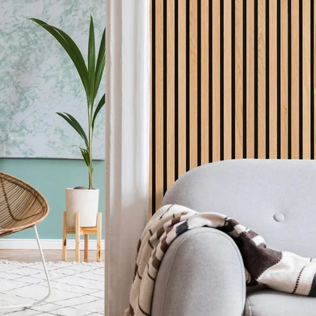A cosy living room corner with a light grey sofa draped with a patterned blanket, a wooden slat accent wall, a green potted plant, and part of a chair with a white frame and woven seat.