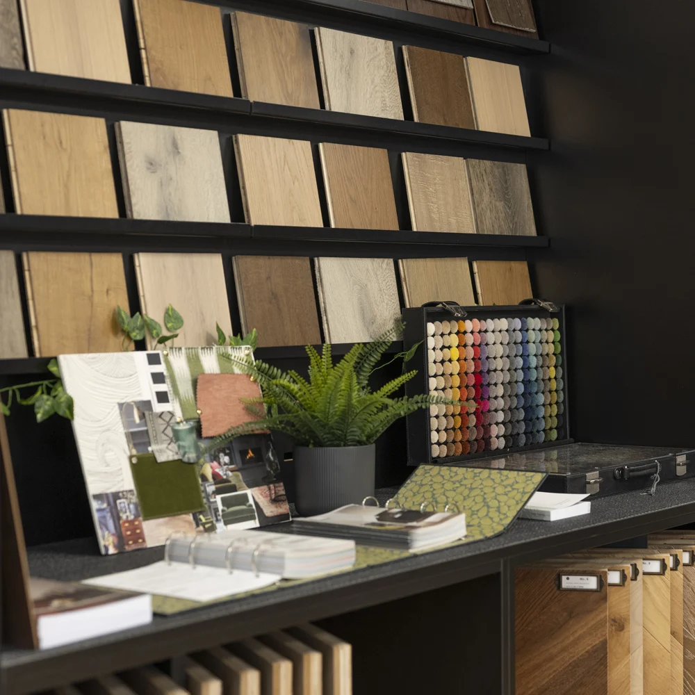 Flooring design studio in Chelmsford. A display of various wood samples on shelves above a counter with fabric swatches, a colour sample case, notebooks, a potted fern, and design materials arranged neatly.