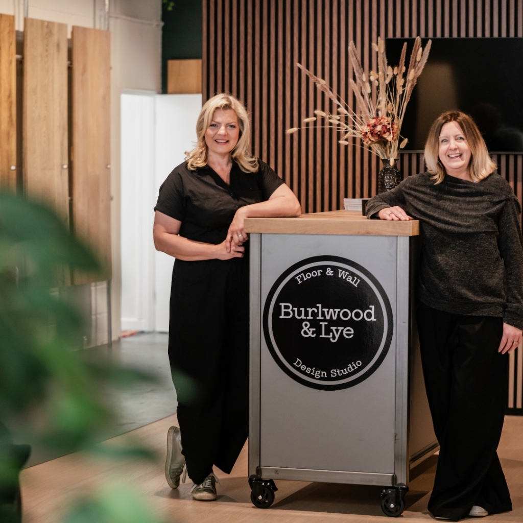Bespoke Curtains and Blinds. Two women stand and lean against a counter with a Burlwood & Lye Design Studio sign in a modern interior featuring wood panelling and a decorative vase with dried plants.
