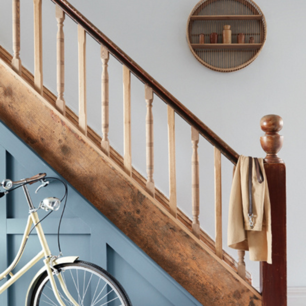 A cream-coloured bicycle leans against a blue panelled wall beneath a wooden staircase. A tan coat and scarf hang from the stair railing. On the wall above, there is a round shelf with small items displayed.