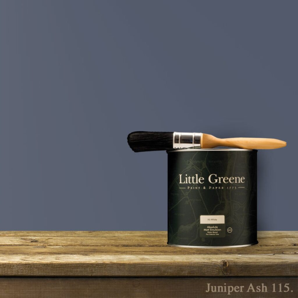A tin of Little Greene paint labelled Juniper Ash 115 sits on a wooden surface with a paintbrush resting across the top. The background wall is painted in a muted, slate blue colour.