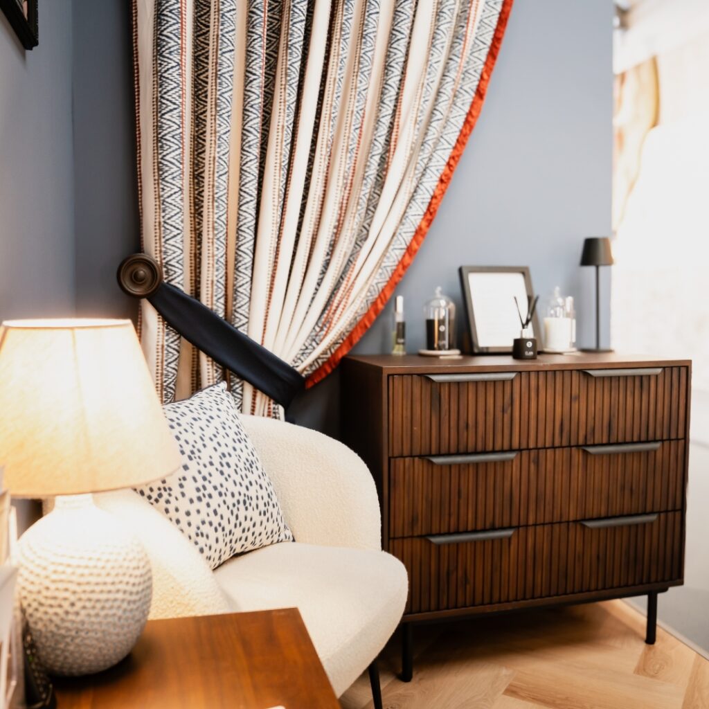 Bespoke Curtains and Blinds. A cosy corner with a white armchair and a wooden chest of drawers, featuring a patterned lamp, decorative cushion, framed photos, and bottles. A large curtain with geometric patterns hangs beside the chest of drawers.