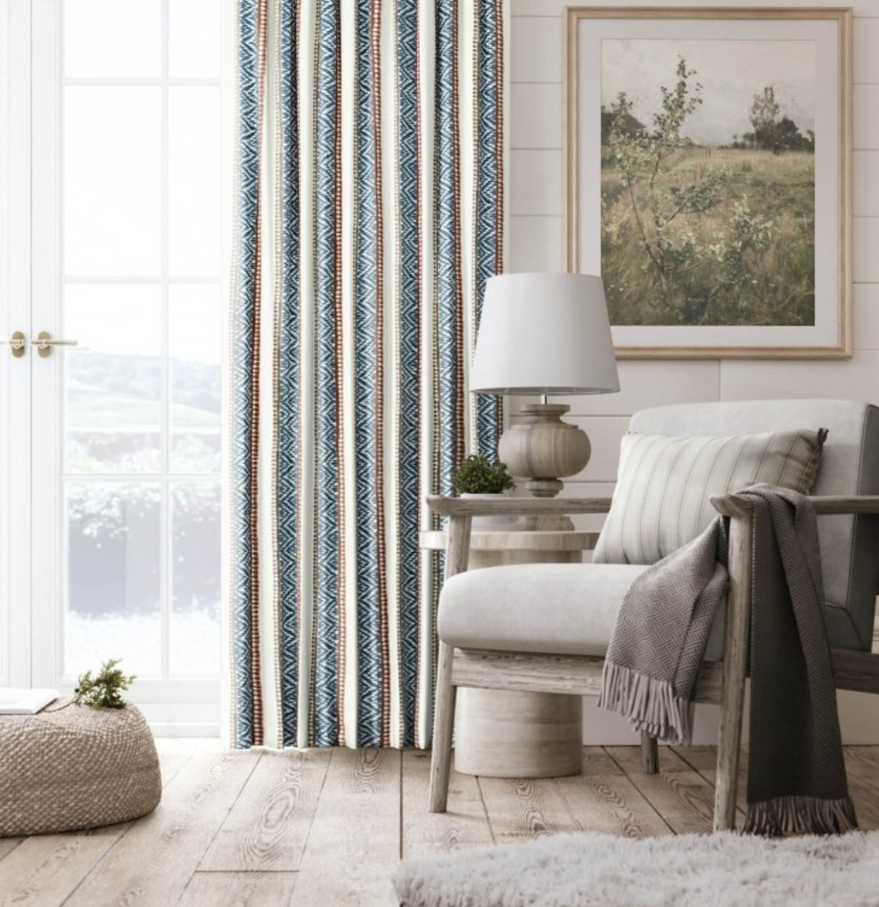 A cosy living room corner with a grey armchair, striped cushion, grey throw blanket, round side table with a lamp, pouffe, patterned curtains, and a framed landscape painting on a white shiplap wall.