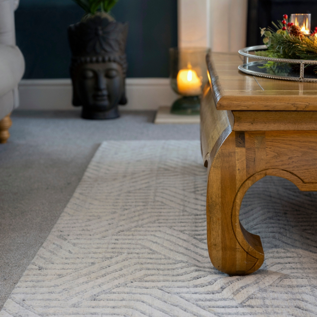 Living Room Rug Ideas. A wooden coffee table sits on a patterned rug in a cosy living room. A candle and greenery decorate the table. In the background, a Buddha head sculpture and another lit candle are visible near the wall.