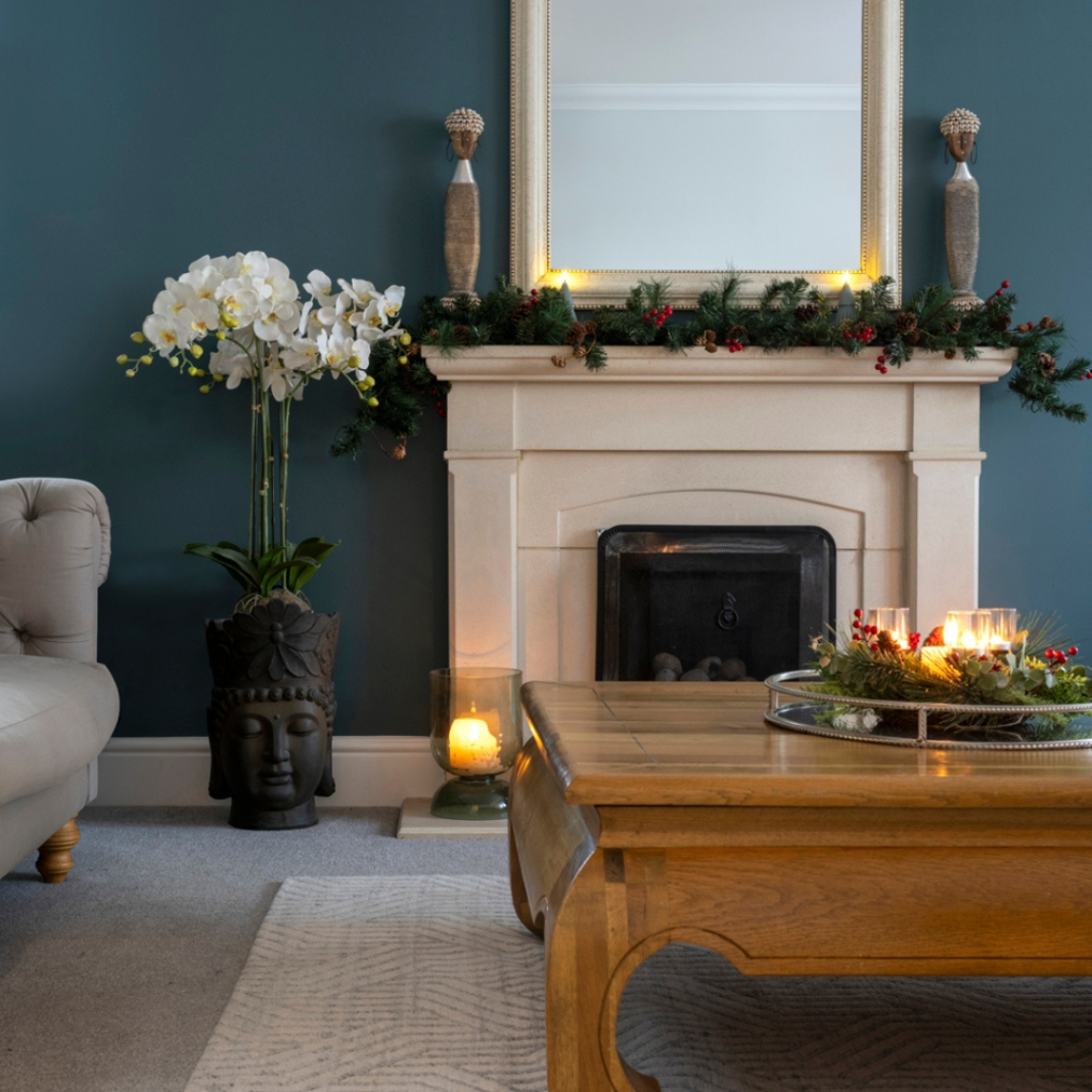Living Room Rug Ideas. A cosy living room with a stone fireplace decorated with garland and candles, a large mirror above it, an orchid plant in a black pot beside a Buddha head, and a wooden coffee table with candles.