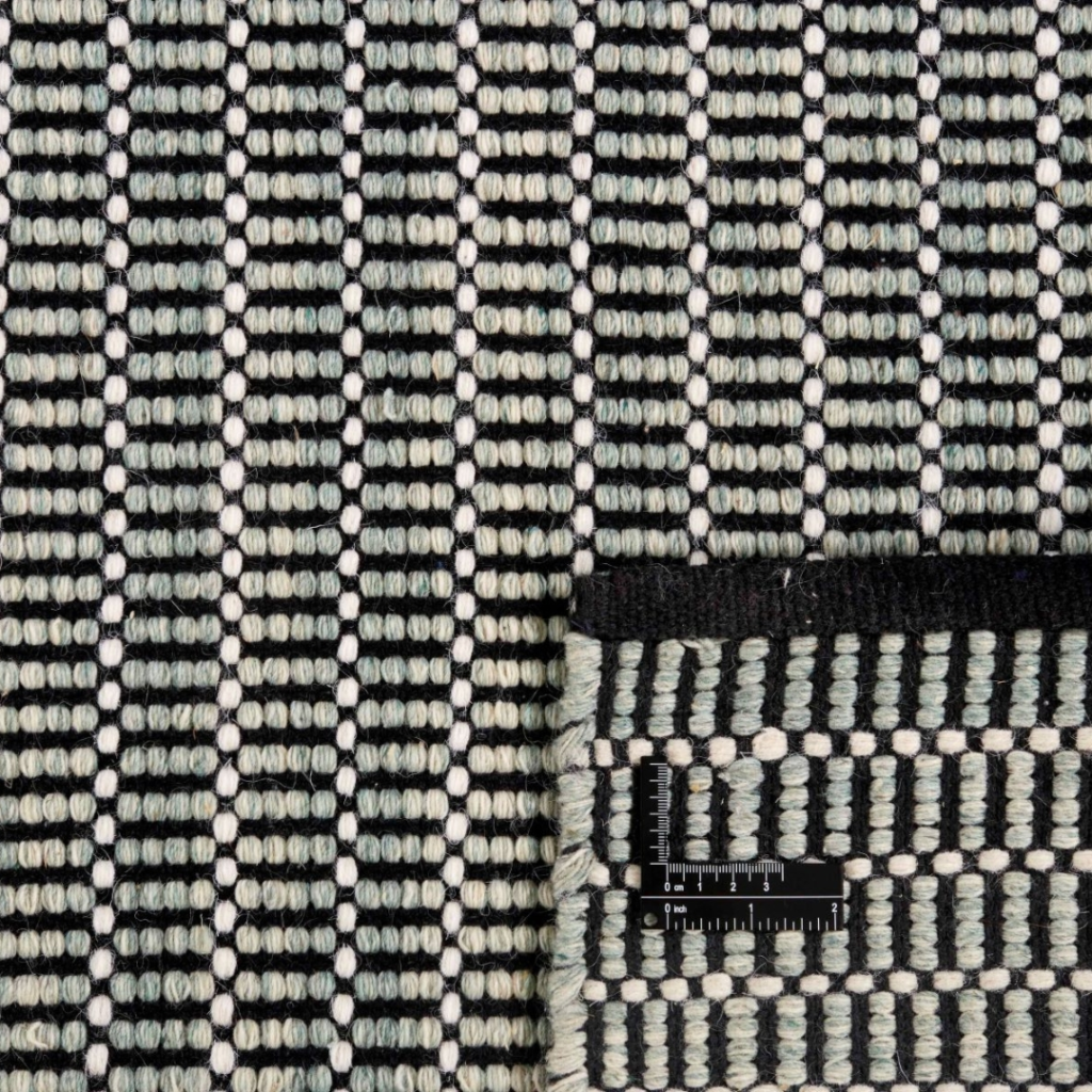 Dining Room Rug Ideas. A close-up of a woven fabric with a geometric pattern of black, white, and grey rows. The bottom right corner is folded, revealing the underside. A small ruler is placed near the fold for scale.