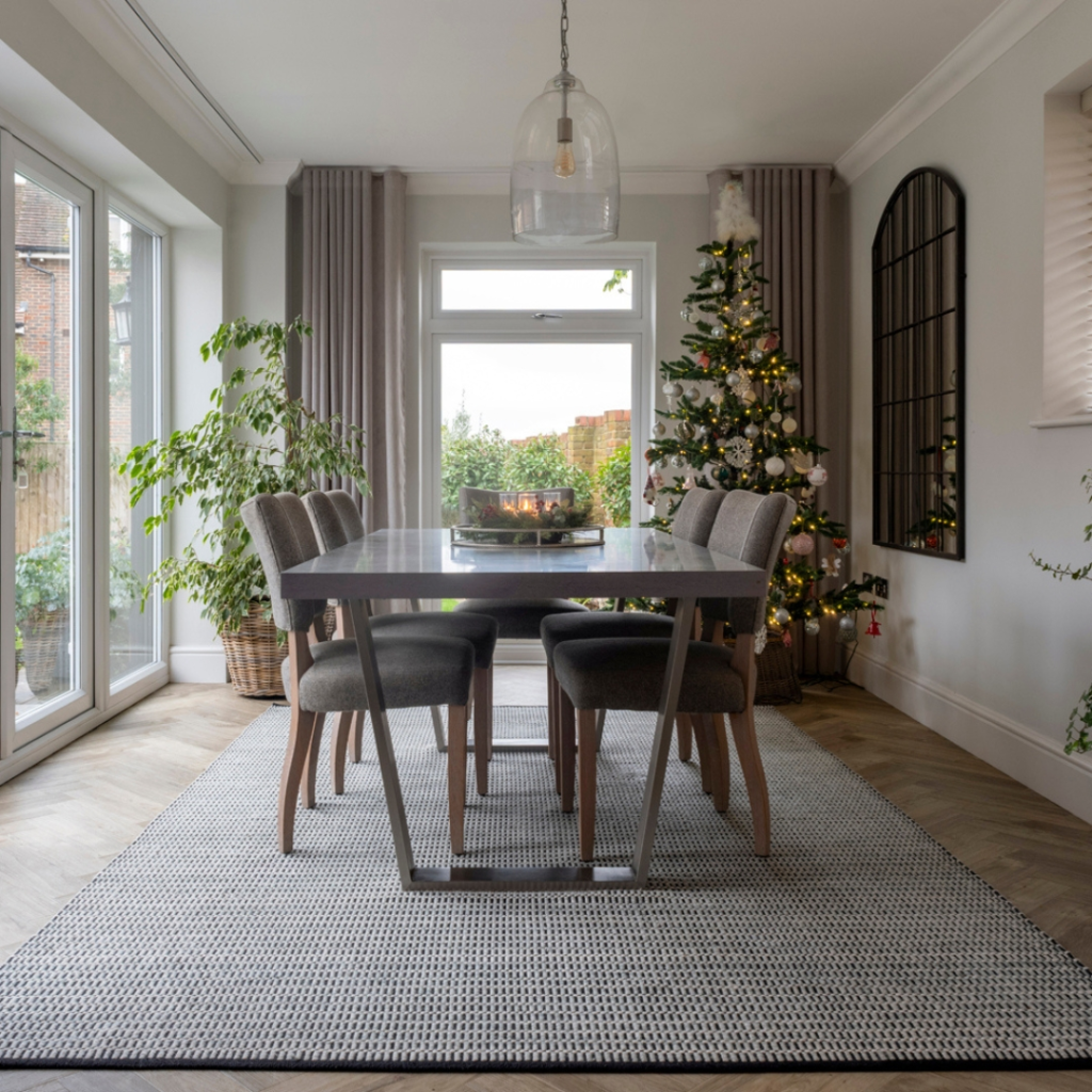 Dining Room Rug Ideas. A modern dining room with a table and six chairs, decorated for Christmas with a lit tree by the window, potted plants, large windows, and a glass pendant light overhead.