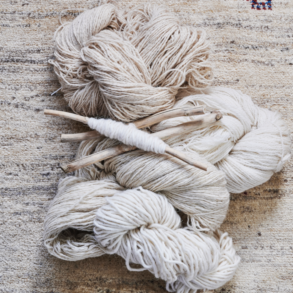 Three skeins of undyed, natural wool yarn and wooden spindles rest on a textured beige rug. One spindle has yarn wound round it, while the others are bare.