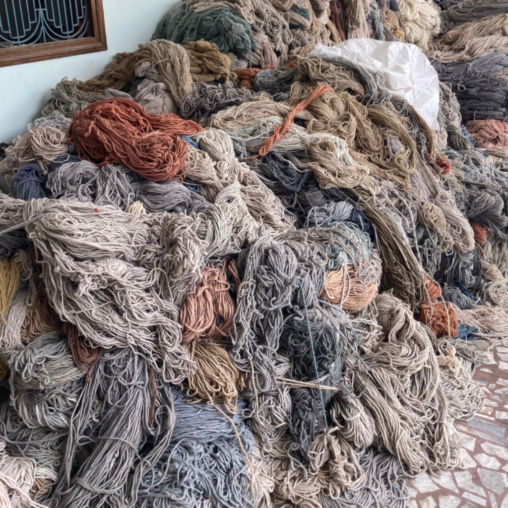 A large pile of tangled, multicoloured yarn is stacked on a tiled floor against a wall, with various shades of beige, brown, grey, and blue threads mixed together.