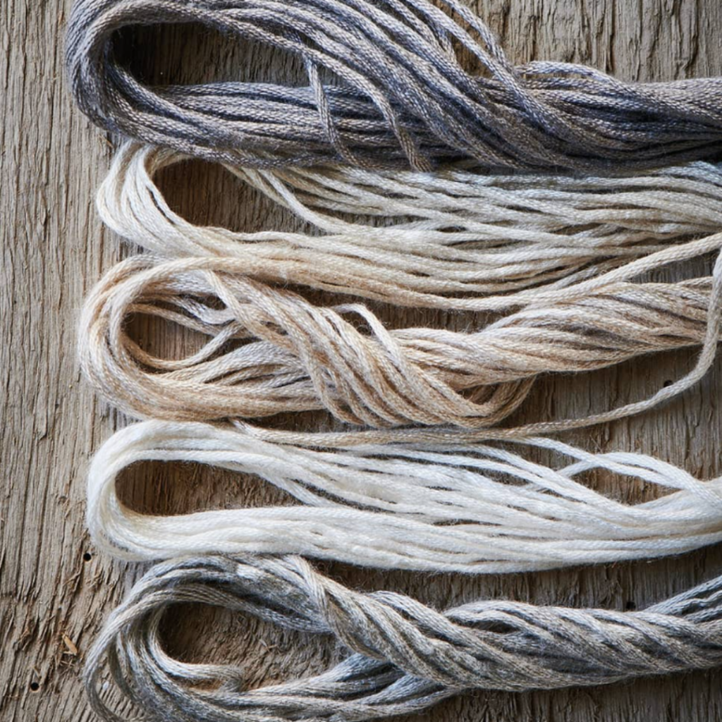 Several bundles of yarn in various natural shades of grey, beige, and white are laid side by side on a textured wooden surface. The yarn is loosely twisted, showcasing its soft and fibrous texture.